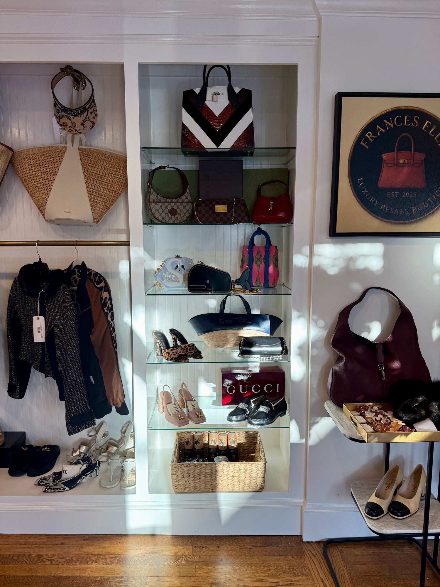 A boutique interior display featuring multiple handbags, shoes, and accessories arranged on glass shelves and racks against a white paneled wall. Visible items include a Gucci box and Gucci-branded bag, a striped tote, woven handbags, and several heels.